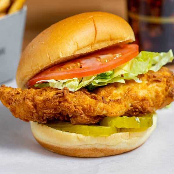 Crispy fried chicken sandwich with lettuce, tomato, and pickles on a bun, with fries and soda in background.