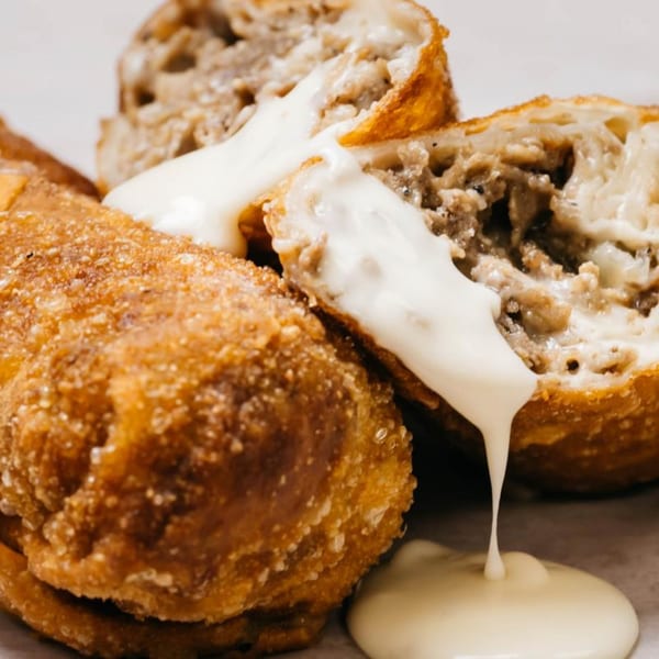 Golden-fried cheesesteak egg rolls, one cut open, with creamy white cheese sauce drizzling over them.