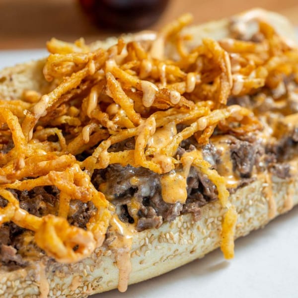 Ochinilis Steaks cheesesteak with crispy fried onions and a creamy sauce on a sesame seed bun.